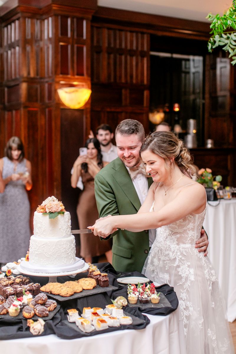 a couple at their wedding reception cutting their wedding cake the woman is wearing a white dress the man is wearing a green suit next to the cake are different desserts and behind them are their wedding guests 