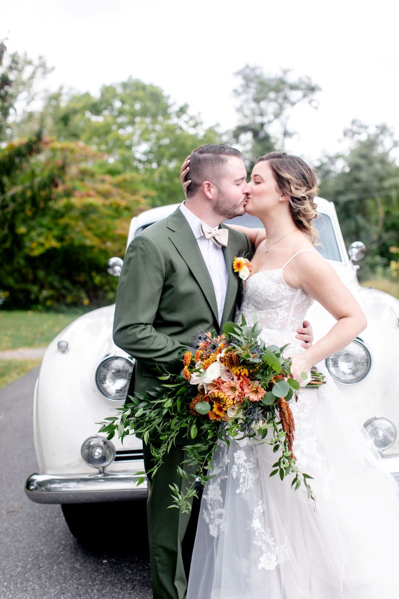 a couple kissing each other in front of a old white car the woman is waering a white wedding dress the man is wearing a green suit and they are holding a bouquet of flowers 