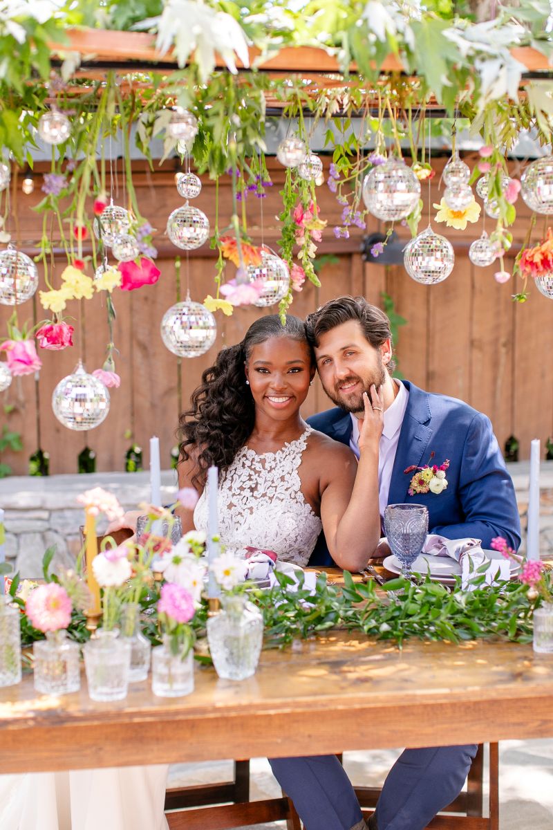 a cuople sitting together at a wooden table the woman is touching the man's face they are both smiling handing over their heads are discoballs and flowers and in front of them are vases with flowers and plates and glasses