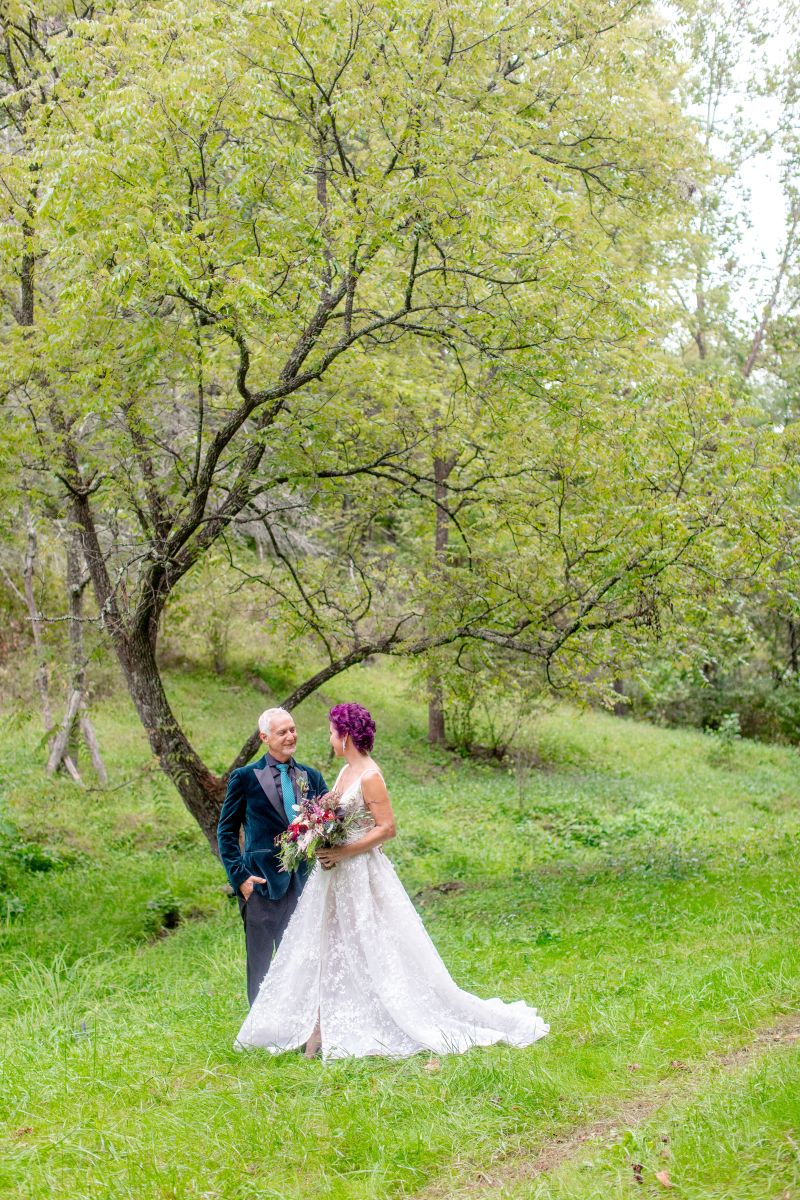 a couple in a green field together looking at one another the woman is waering a white wedding dress and holding a bouquet of flowers the man is wearing a suit they are surrounded by trees and nature 