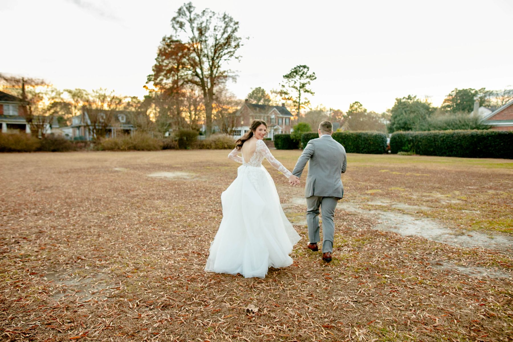 a couple walking across a lawn together holding hands the man is wearing a gray suit and the woman is wearing a white wedding dress and is looking over her shoulder 