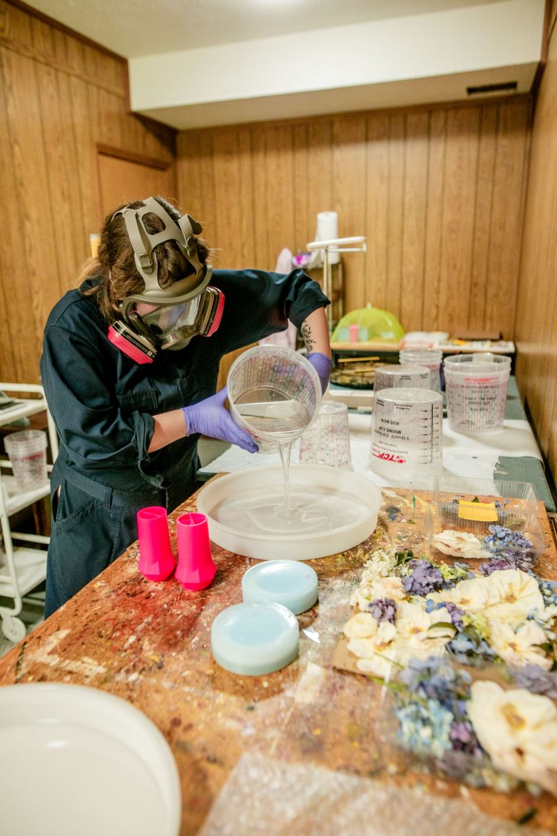 a woman wearing a gas mask pouring a clear liquid into a bowl inside of her worksapce 