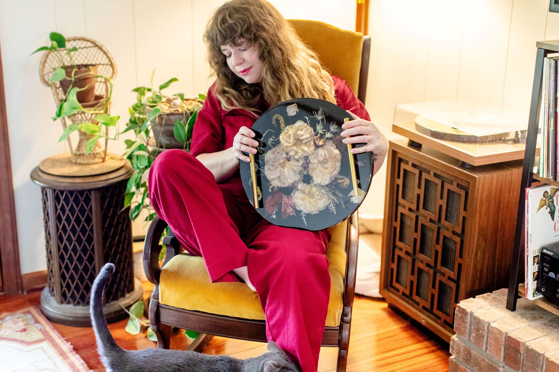 a woman sitting in a chair her cat is bumping into her leg and she is holding a frame with flowers preserved inside 