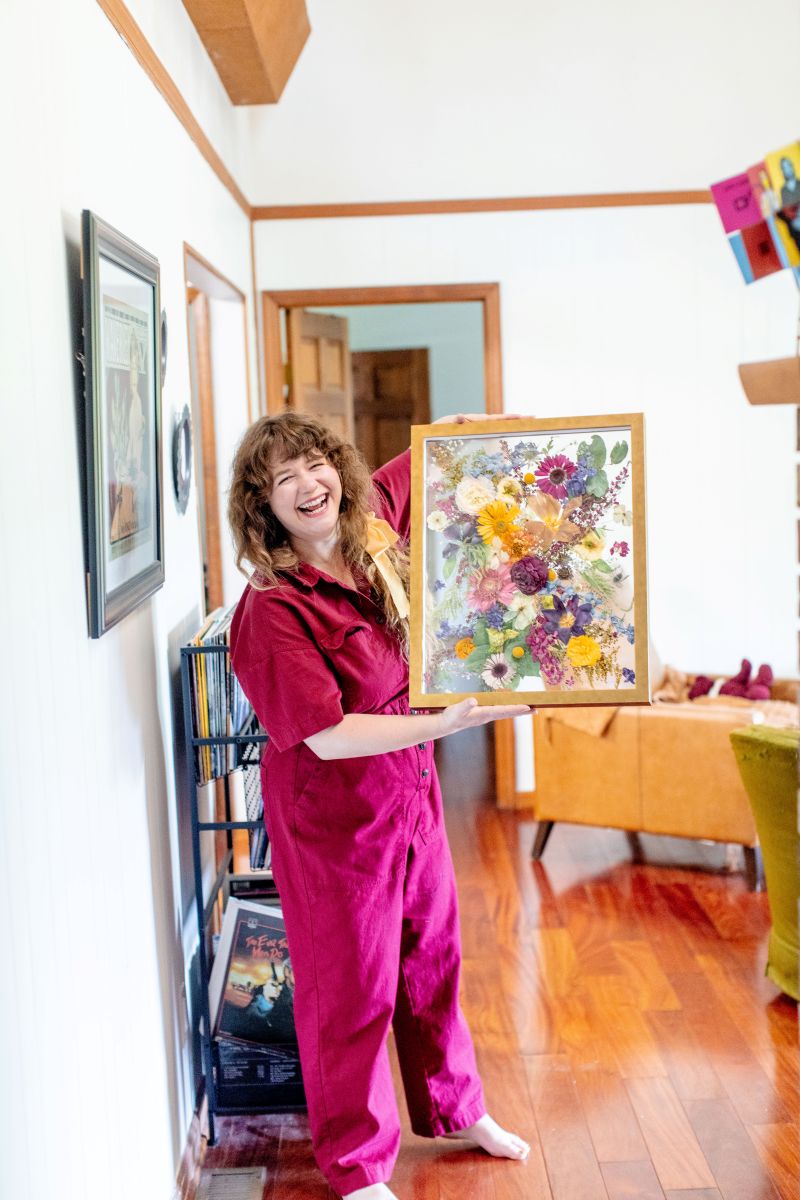 A woman in a red jumper holding up a frame with flowers preserved inside of it 