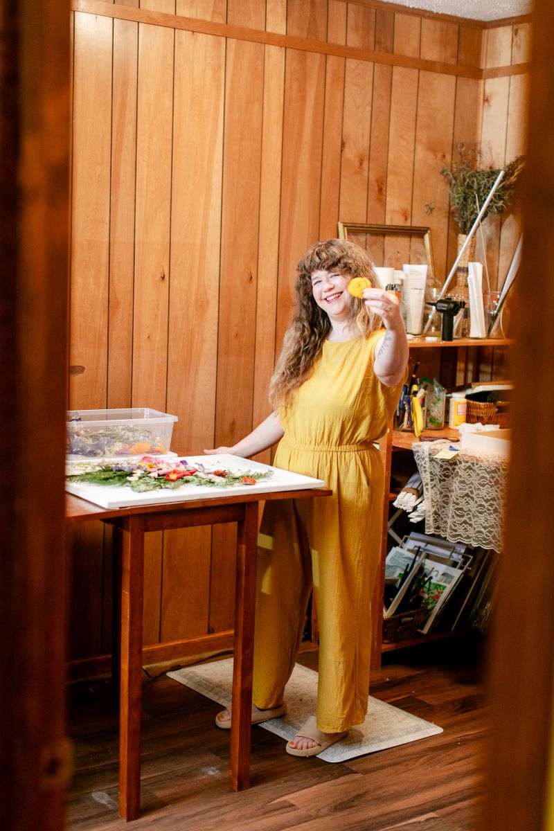 A woman in a yellow jumper in her workspace holding up a yellow flower 