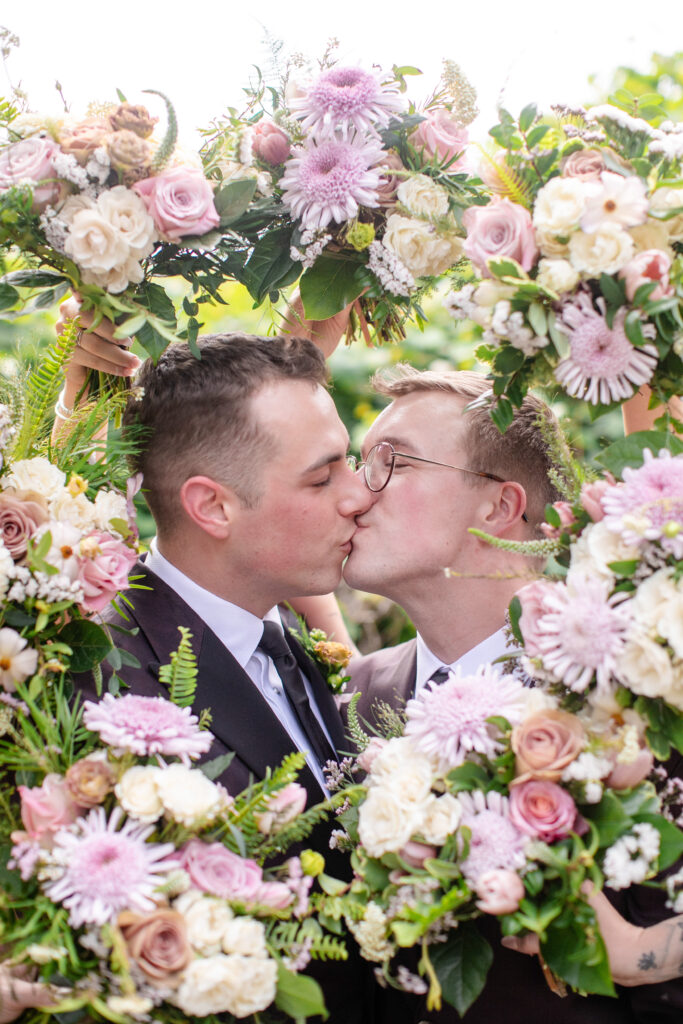 LGBTQ friendly wedding photographer Asheville documenting two grooms sharing a kiss framed by pastel wedding bouquets