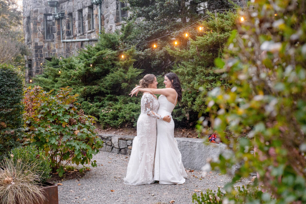 LGBTQ friendly wedding photographer Asheville capturing two brides embracing during their outdoor wedding at Homewood in Asheville with string lights and stone architecture
