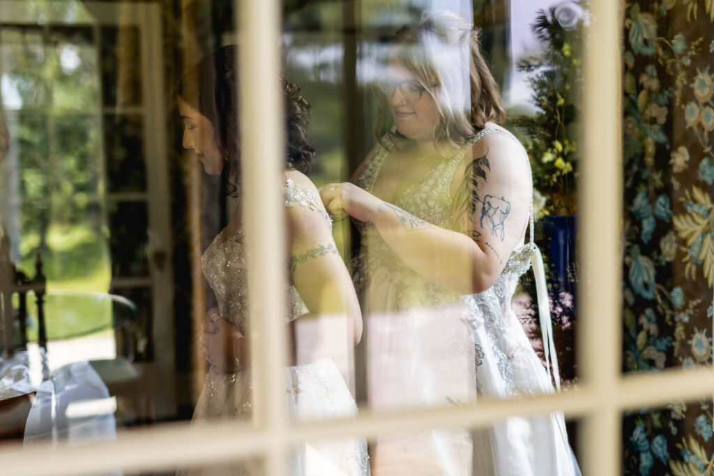 LGBTQ friendly wedding photographer Asheville capturing a bride helping her partner fasten her dress through a window reflection before their ceremony