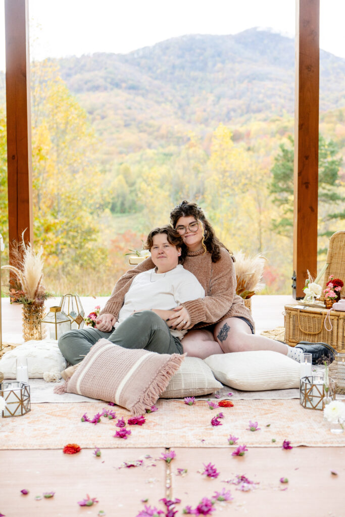 LGBTQ friendly wedding photographer Asheville capturing a mountain proposal with an LGBTQ couple sitting together on a styled picnic setup overlooking the fall Blue Ridge scenery
