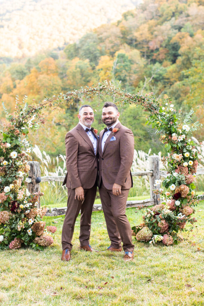LGBTQ friendly wedding photographer Asheville capturing two grooms standing together under a floral arch with fall mountain views in the Blue Ridge Mountains
