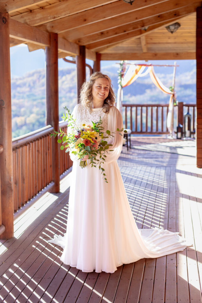 Bride standing on a cabin deck during her Asheville elopement with the Blue Ridge Mountains in the background holding a colorful bouquet