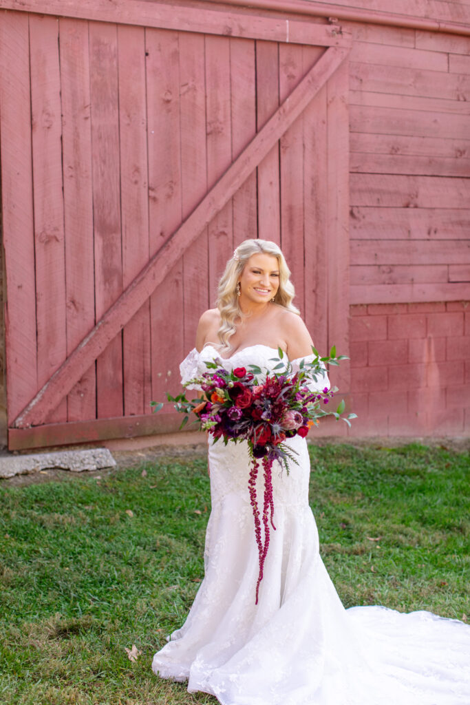 Bride holding a deep burgundy and blush bouquet in front of the red barn at Honeysuckle Hill, wearing an off the shoulder lace wedding gown during portraits.