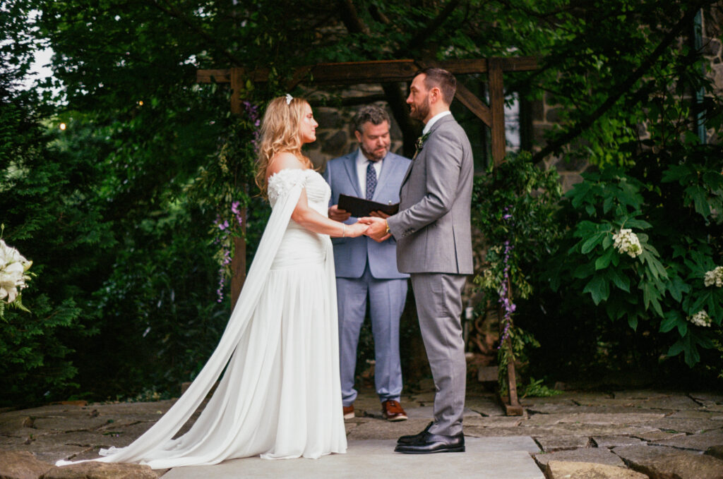 Bride and groom exchanging vows during an intimate outdoor ceremony at Homewood in Asheville, photographed on film beneath a wooden arbor and surrounding greenery.