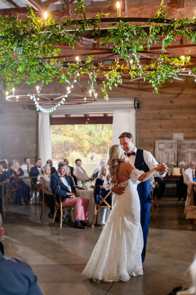 Bride and groom share their first dance during a Honeysuckle Hill wedding reception, surrounded by guests beneath a greenery-covered chandelier in the barn.