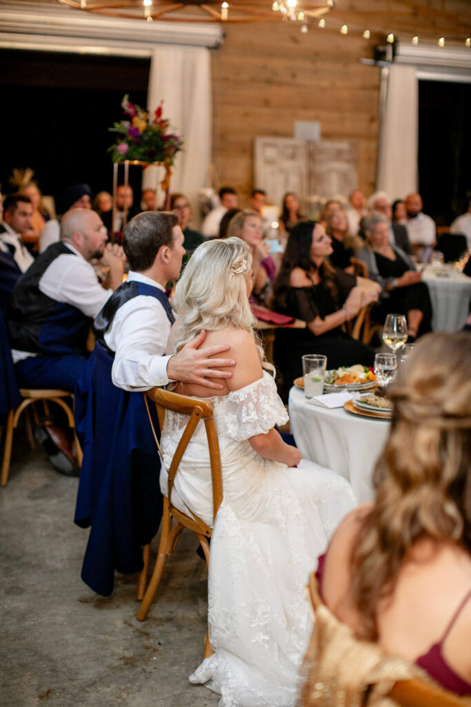 Bride and groom seated together during their wedding reception at Honeysuckle Hill, listening to speeches surrounded by guests in a warmly lit barn setting.