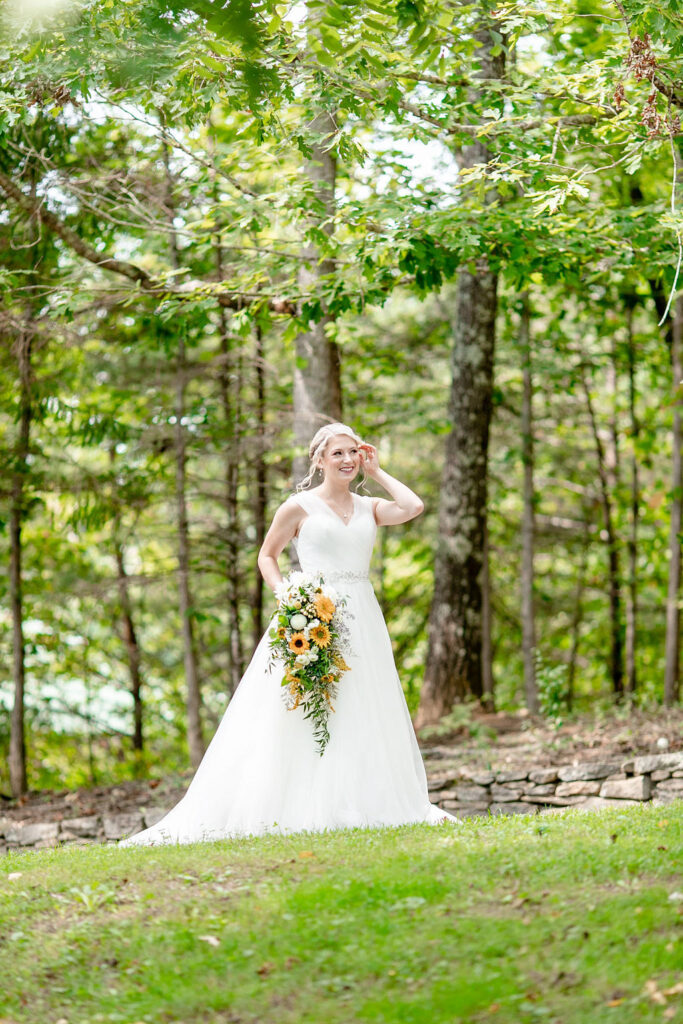 Bride holding a sunflower bouquet standing in a wooded clearing at Honeysuckle Hill, wearing a flowing white wedding dress surrounded by lush green trees.
