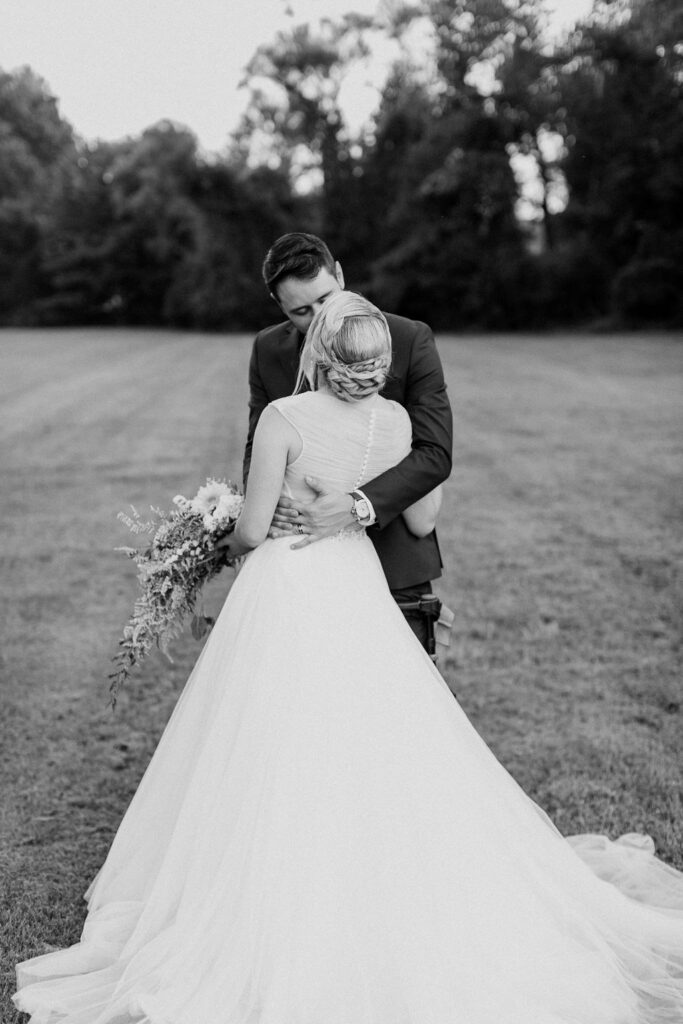 Black and white portrait of a bride and groom embracing during golden hour at Honeysuckle Hill wedding venue in Asheville, surrounded by open fields and trees