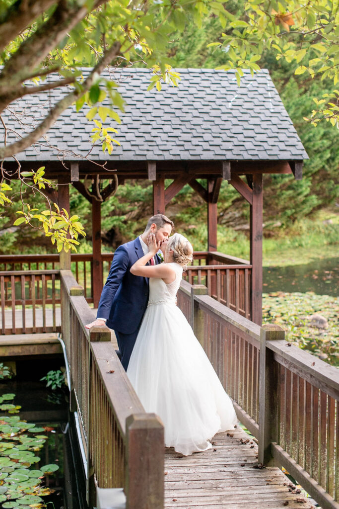 Bride and groom sharing a kiss on a wooden bridge over the pond at Honeysuckle Hill, surrounded by trees and greenery during golden hour.