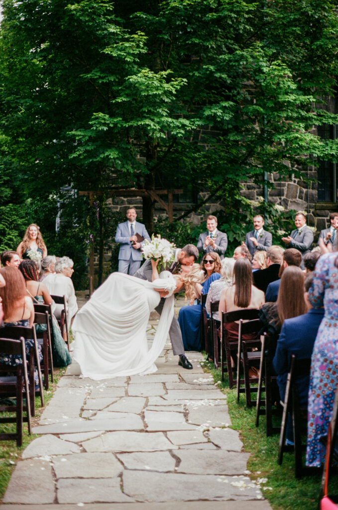 Bride and groom share a celebratory kiss in the aisle during an outdoor wedding ceremony at Homewood in Asheville, surrounded by guests and lush green trees.