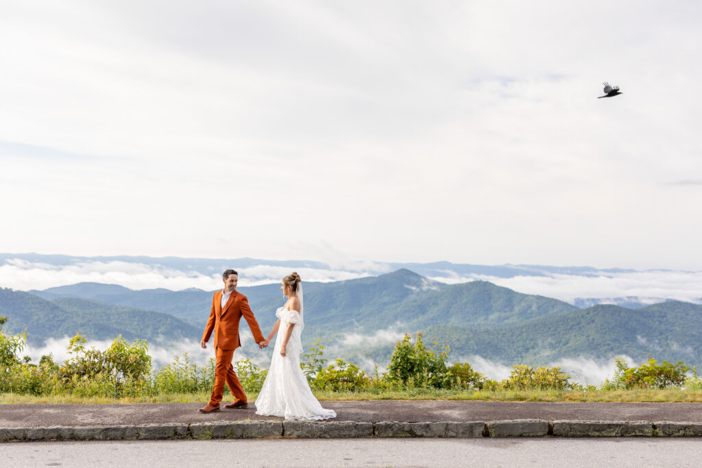 bride and groom holding hands walking along the Blue Ridge Parkway overlook with foggy mountain views on their Asheville NC wedding day
