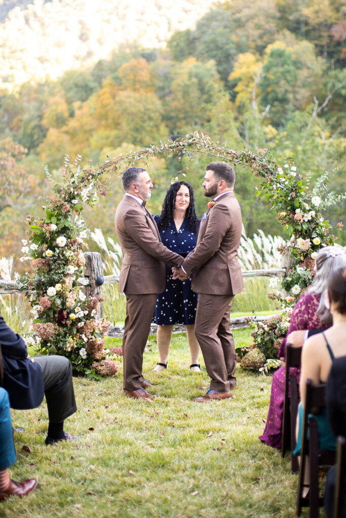  two grooms exchanging vows under a floral arch at an outdoor fall wedding ceremony in Asheville NC