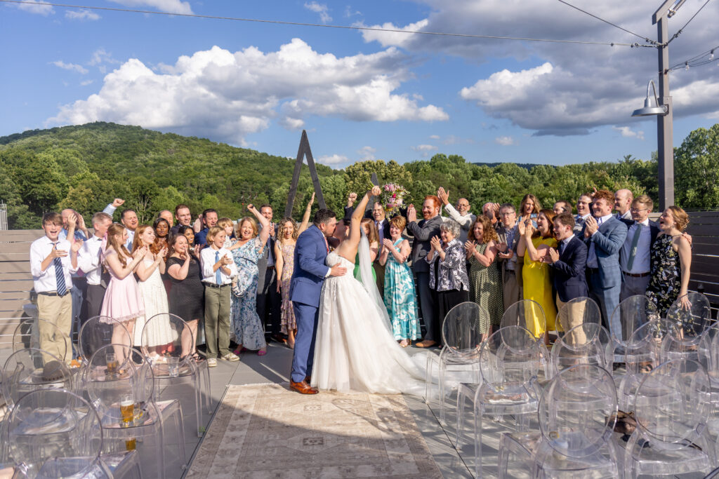 bride and groom first kiss surrounded by cheering guests at rooftop wedding ceremony at Highland Brewing Company in Asheville NC