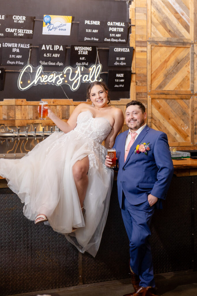 bride and groom holding Highland Brewing beers at the bar with cheers y'all neon sign at Highland Brewing Company wedding in Asheville NC