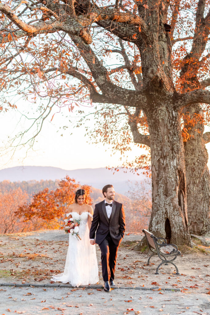 bride and groom walking hand in hand under a large oak tree with fall foliage and Blue Ridge Mountain views at an Asheville NC wedding